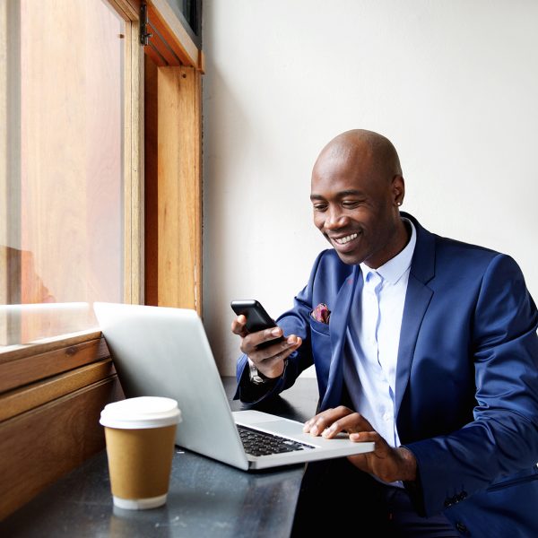 Portrait of happy businessman using phone while working on laptop in a restaurant.