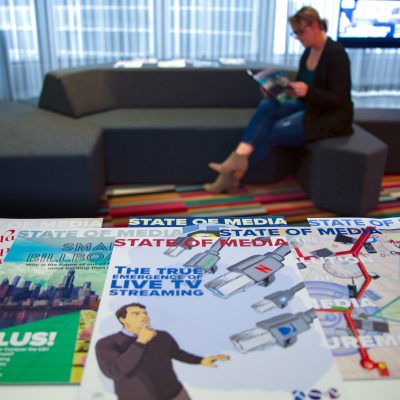 Image of a woman sitting on a couch and reading a magazine with a stack of magazines in front of her.