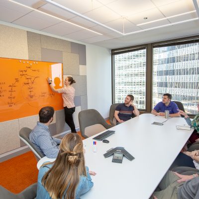 Image of a group of people sitting around a table watching a woman write on a dry erase board.